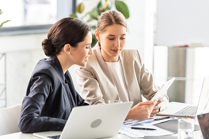 Two businesswomen collaborating at a desk with laptops and documents in a bright modern office.