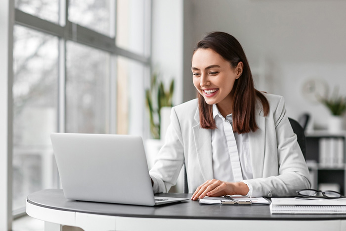 Businesswoman in white blazer smiling while working on laptop at modern office desk.